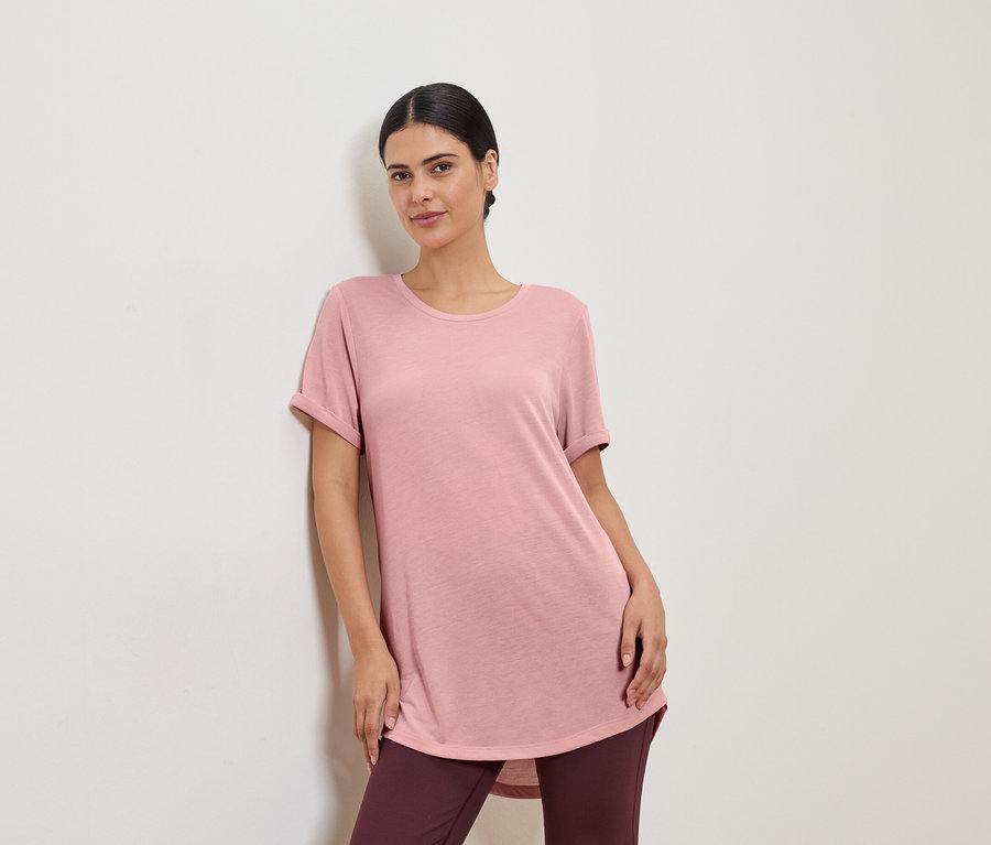 Frau in rosafarbenem T-Shirt und Leggings, die an einer weißen Wand lehnt.