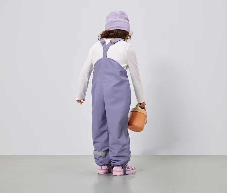 Rückansicht eines Kindes mit lila Mütze und Overall, weißem Langarmshirt, rosa Schuhen und einem orangefarbenen Eimer.