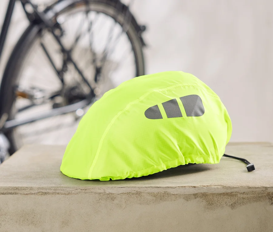 Ein neongelber Fahrradhelmüberzug mit reflektierenden Streifen liegt auf einem Betonsockel, im Hintergrund ein Fahrrad.