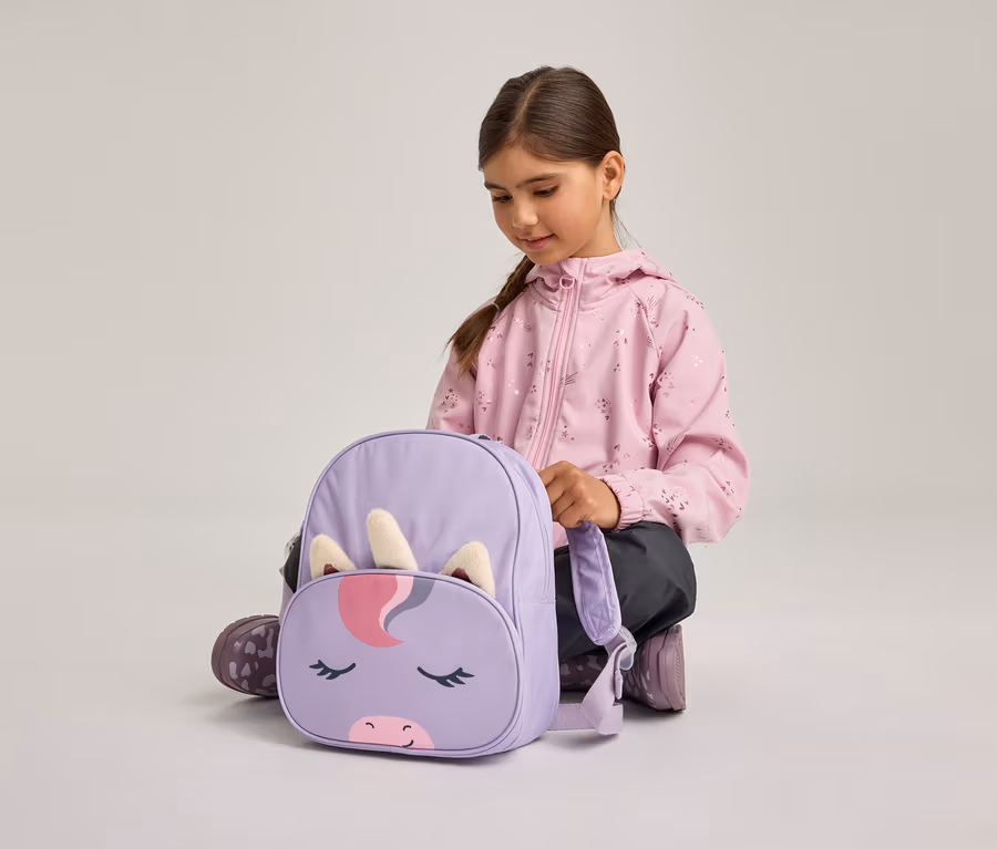 Ein junges Mädchen in einer rosa Jacke und dunklen Hosen hockt am Boden und blickt auf einen lila Rucksack mit Einhorn-Gesichtsdesign, inklusive Horn und Ohren.