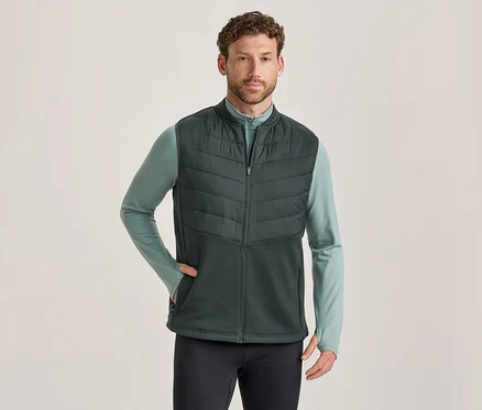 Mann trägt eine dunkelgrüne Steppweste über einem hellgrünen Langarmshirt und dunklen Hosen, eine Hand in der Westentasche.