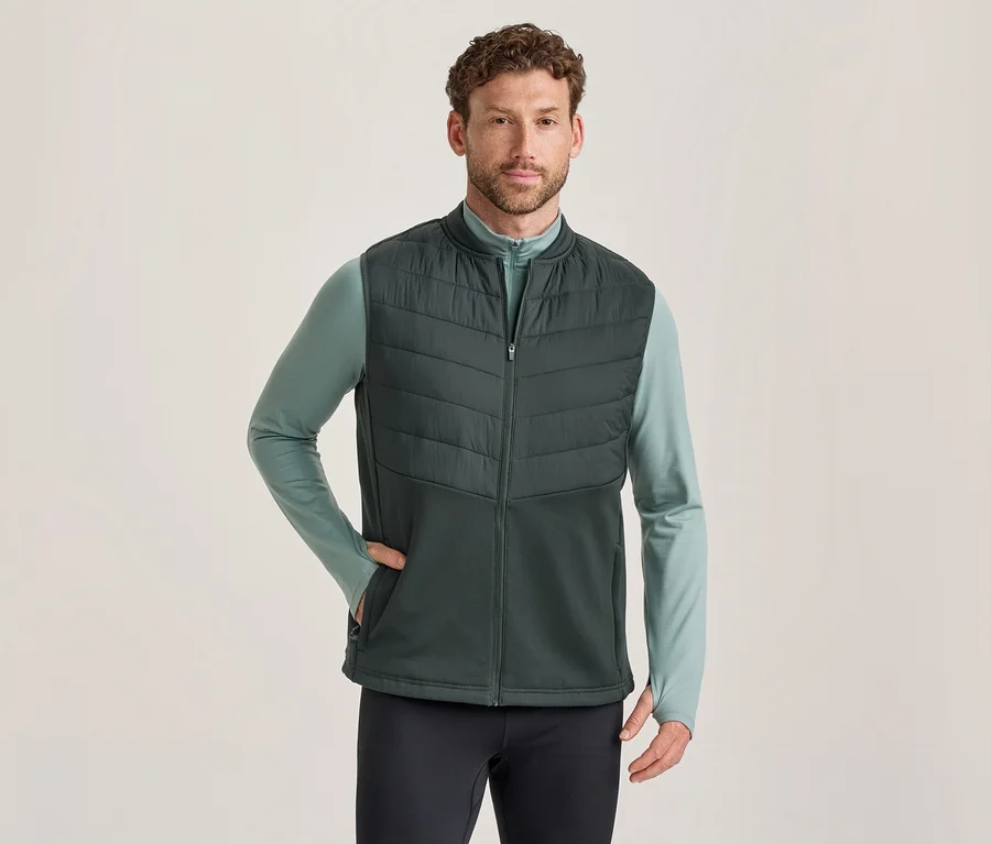 Mann trägt eine dunkelgrüne Steppweste über einem hellgrünen Langarmshirt und dunklen Hosen, eine Hand in der Westentasche.