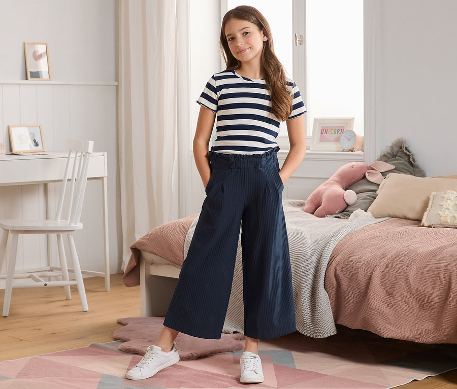 Ein Mädchen posiert in einem Zimmer und trägt eine plissierte Culotte mit recyceltem Material und ein gestreiftes T-Shirt.