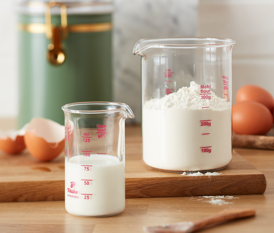 Zwei Messbecher, einer mit Milch und einer mit Mehl, stehen neben Eiern auf einem Holztisch.