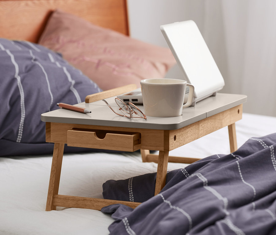 Auf einem Bett steht ein Betttisch mit einem Laptop, einer Tasse und einer Brille.