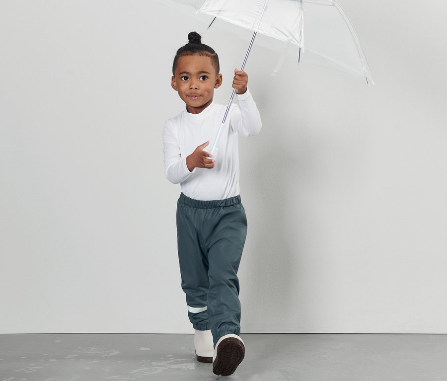 Ein Junge hält einen durchsichtigen Regenschirm und trägt eine Kleinkind-Regenhose.