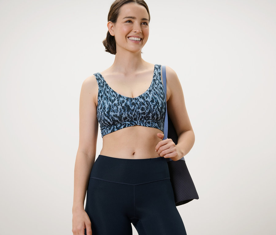 Frau trägt ein Sport-Bustier mit Alloverprint und hält eine Trainingsmatte.