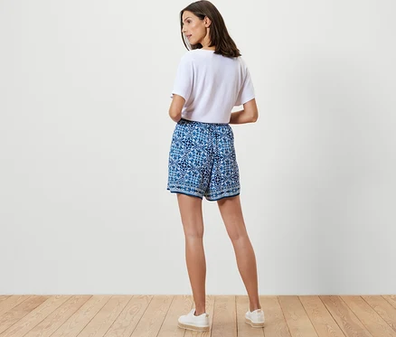 Frau steht mit dem Rücken zur Kamera und trägt bedruckte Webshorts und ein strukturiertes, weißes Shirt.