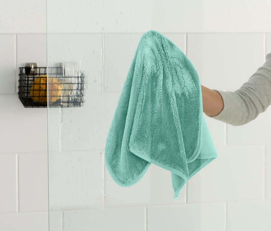 Eine Person wischt eine gläserne Duschwand mit einem türkisfarbenen Handtuch ab. In der Ecke befindet sich ein schwarzer Drahtkorb.