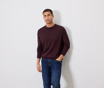 Mann posiert vor einem weißen Hintergrund und trägt einen burgunderfarbenen Pullover und Jeans.