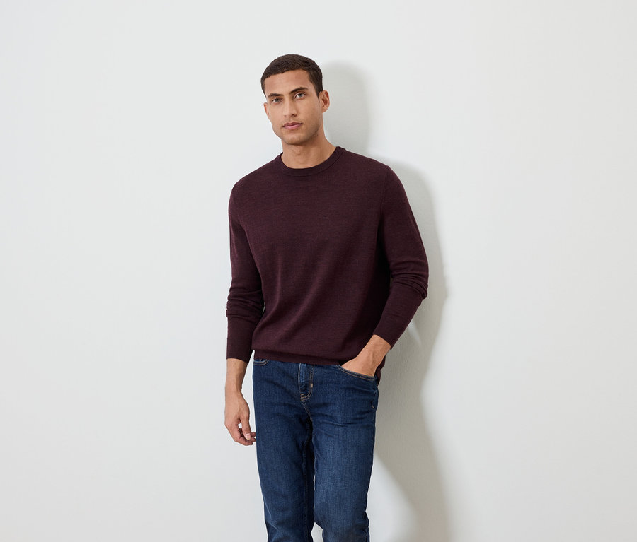 Mann posiert vor einem weißen Hintergrund und trägt einen burgunderfarbenen Pullover und Jeans.
