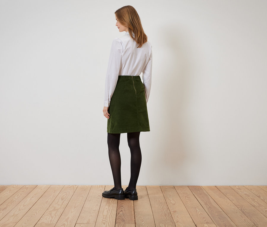 Eine Frau steht mit dem Rücken zur Kamera und trägt einen grünen Cordrock, ein weißes Hemd, eine schwarze Strumpfhose und schwarze Schuhe.