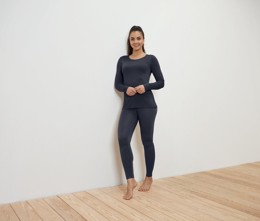 Frau in dunkelblauer Thermounterwäsche steht an einer weißen Wand auf einem Holzboden.