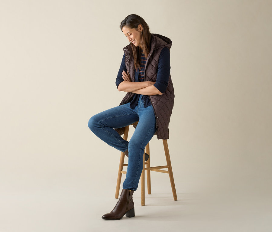 Eine Frau sitzt mit verschränkten Armen auf einem Hocker und trägt eine lange Steppweste mit Kapuze in Dunkelbraun.