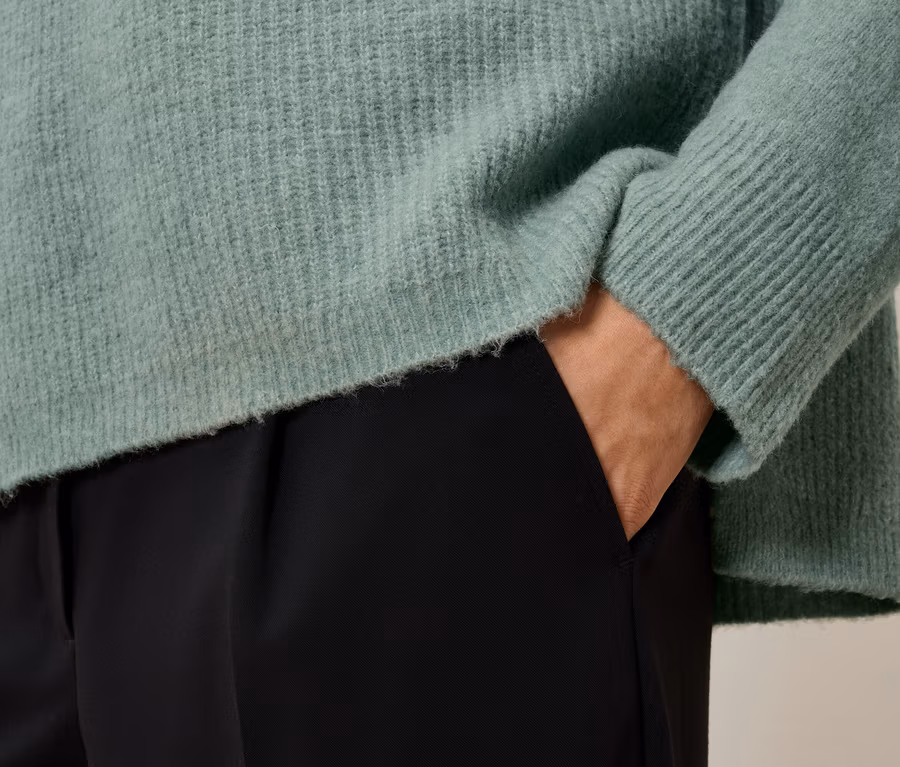 Detailaufnahme eines Damenoutfits: Ein hellgrüner Pullover und eine schwarze Hose. Eine Hand steckt in der Hosentasche.
