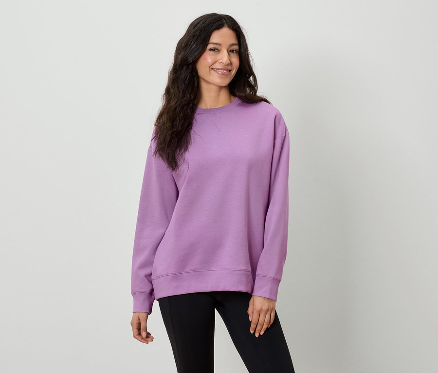 Eine Frau mit langen braunen Haaren trägt ein lila Sweatshirt und eine schwarze Hose.