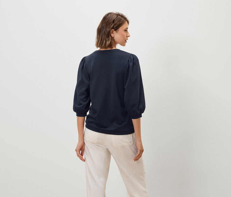 Eine Frau trägt ein dunkelblaues Oberteil mit Dreiviertelärmeln und eine beige Hose.