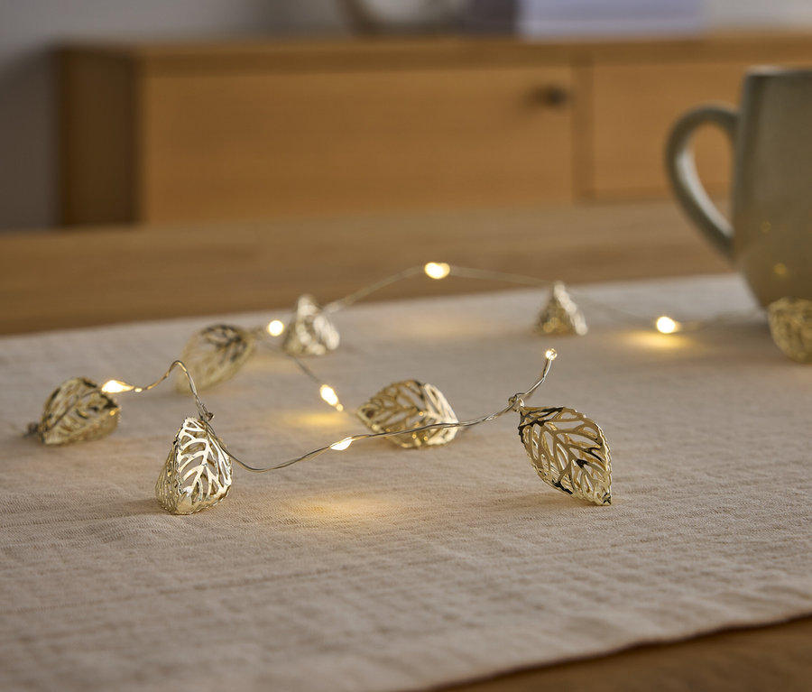 Eine goldene Lichterkette in Blattform liegt auf einem Tischläufer neben einer Tasse.