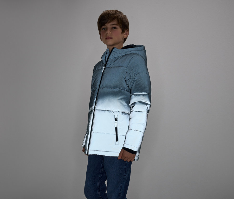 Junge in blauem Steppjacke und Jeans steht vor grauem Hintergrund.