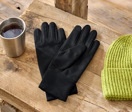Auf einem Holztisch stehen ein Metallbecher, schwarze Handschuhe und eine grüne Strickmütze.