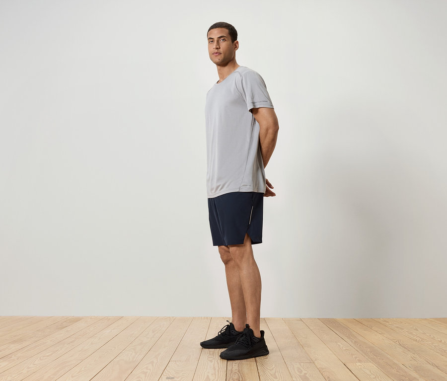 Mann steht seitlich, trägt ein graues T-Shirt, dunkelblaue Shorts und schwarze Turnschuhe.