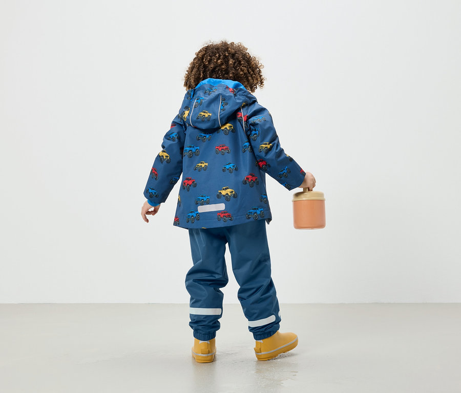 Junge in blauer Jacke mit Autoaufdruck hält einen orangefarbenen Behälter.