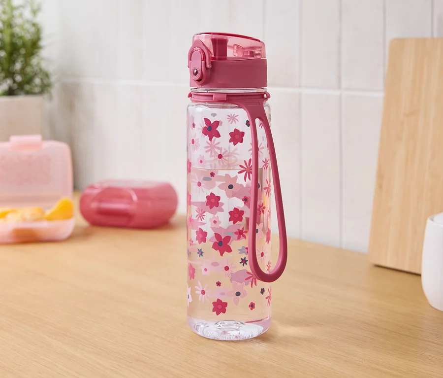 Auf einem Holztisch stehen eine durchsichtige Wasserflasche mit rosa Blüten, rosa Deckel und Schlaufe. Im Hintergrund befinden sich eine Brotdose mit Mandarinen, eine geschlossene Dose, ein Schneidebrett und eine Zuckerdose.