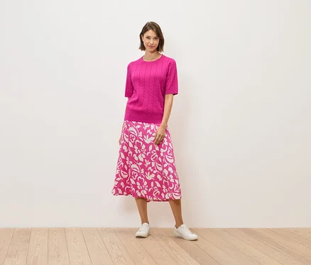 Eine Frau in einem rosa Pullover und einem Rock mit Blumenmuster steht auf einem Holzboden.