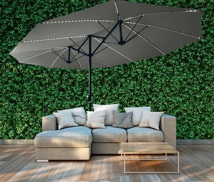Ein grauer LECO Doppel-Sonnenschirm mit LED-Beleuchtung steht über einem Sofa mit Kissen und einem kleinen Holztisch.