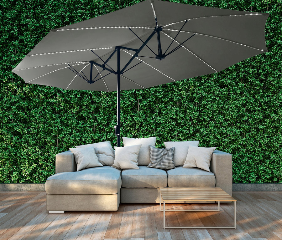 Ein grauer LECO Doppel-Sonnenschirm mit LED-Beleuchtung steht über einem Sofa mit Kissen und einem kleinen Holztisch.