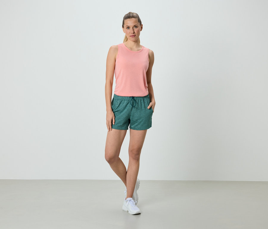Frau steht mit den Händen in den Taschen in grünen Shorts und einem rosa Tanktop.