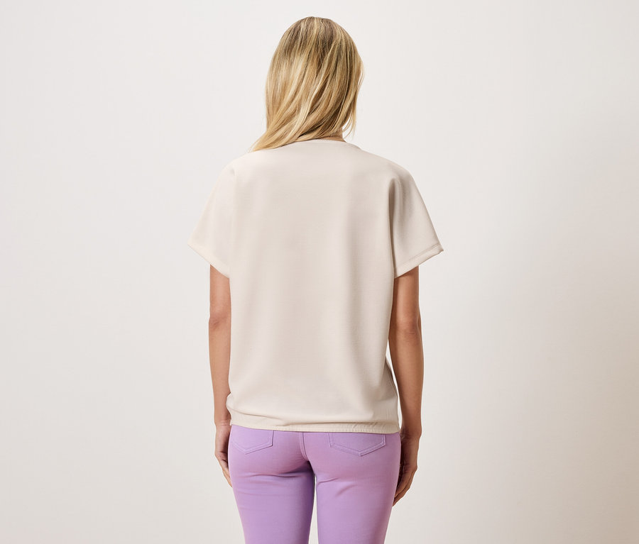 Rückansicht einer Frau mit blonden Haaren, die ein beigefarbenes Oberteil und lila Hosen trägt.