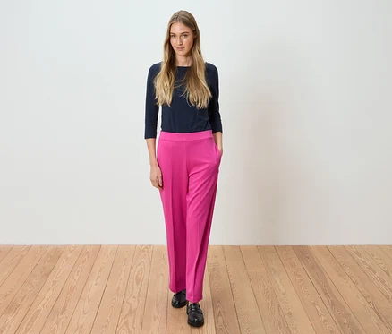 Frau steht in marineblauem Oberteil und pinker Hose vor einer weißen Wand.