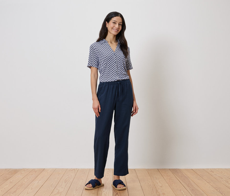 Eine Frau steht vor einer Wand und trägt ein blaues geometrisches Hemd und eine marineblaue Hose.