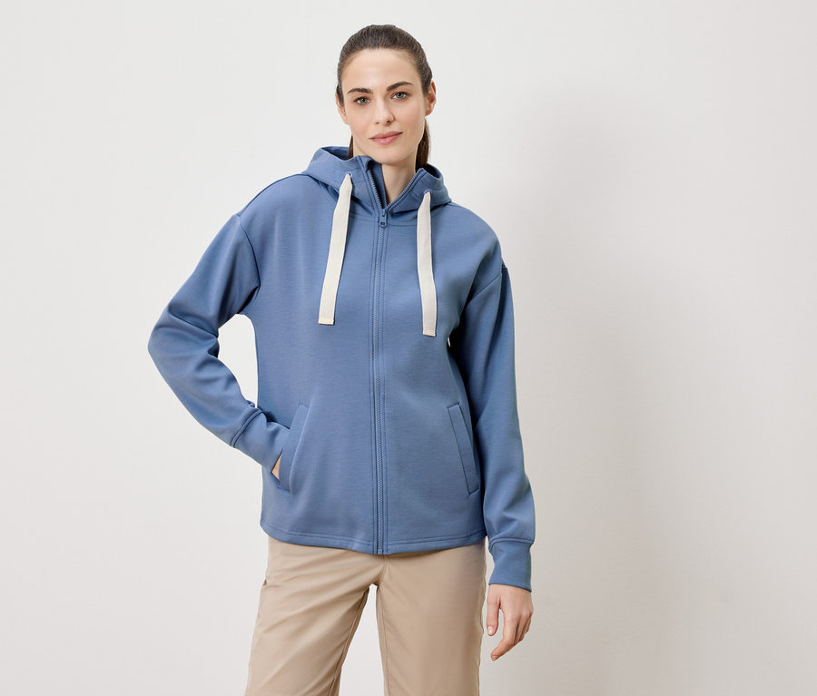 Frau mit Zopf trägt einen blauen Kapuzenpullover mit Reißverschluss und beigen Hosen.