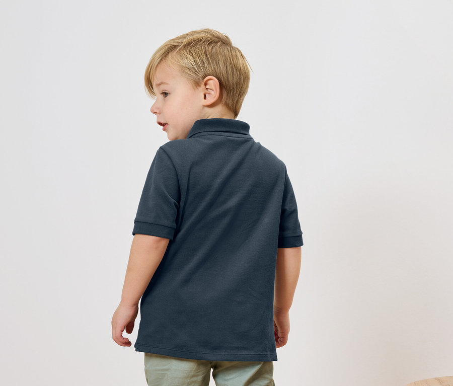 Ein blonder Junge trägt ein dunkelblaues Poloshirt und hellgrüne Hosen, von hinten aufgenommen.