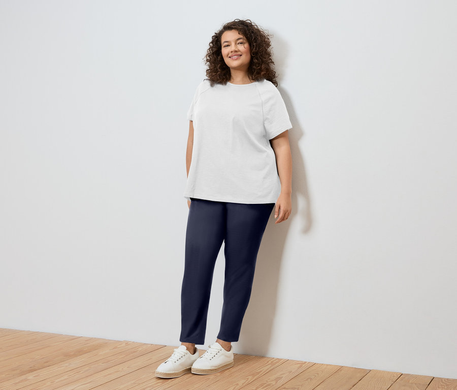 Frau mit lockigem Haar, trägt ein weißes T-Shirt, blaue Hosen und weiße Schuhe, steht an einer weißen Wand.