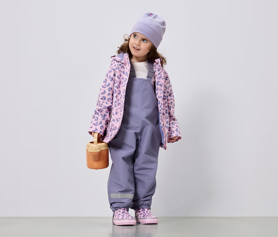 Ein kleines Mädchen mit lockigem Haar trägt einen lila Overall, eine rosa-lila Jacke mit Leopardenmuster, eine Mütze und passende Schuhe. Sie hält eine Gießkanne.