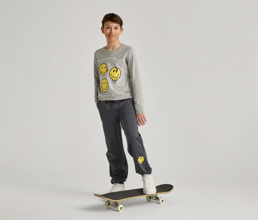 Ein Junge mit grauem Sweatshirt und Hose mit gelben Smileys steht auf einem Skateboard vor einem weißen Hintergrund.