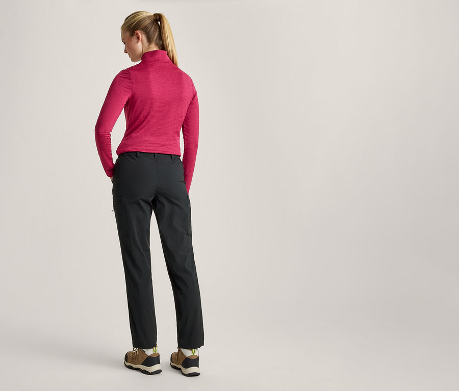 Frau von hinten, in einem pinkfarbenen Langarmshirt, dunkelgrauen Hosen und Wanderschuhen, mit den Händen in den Hosentaschen.