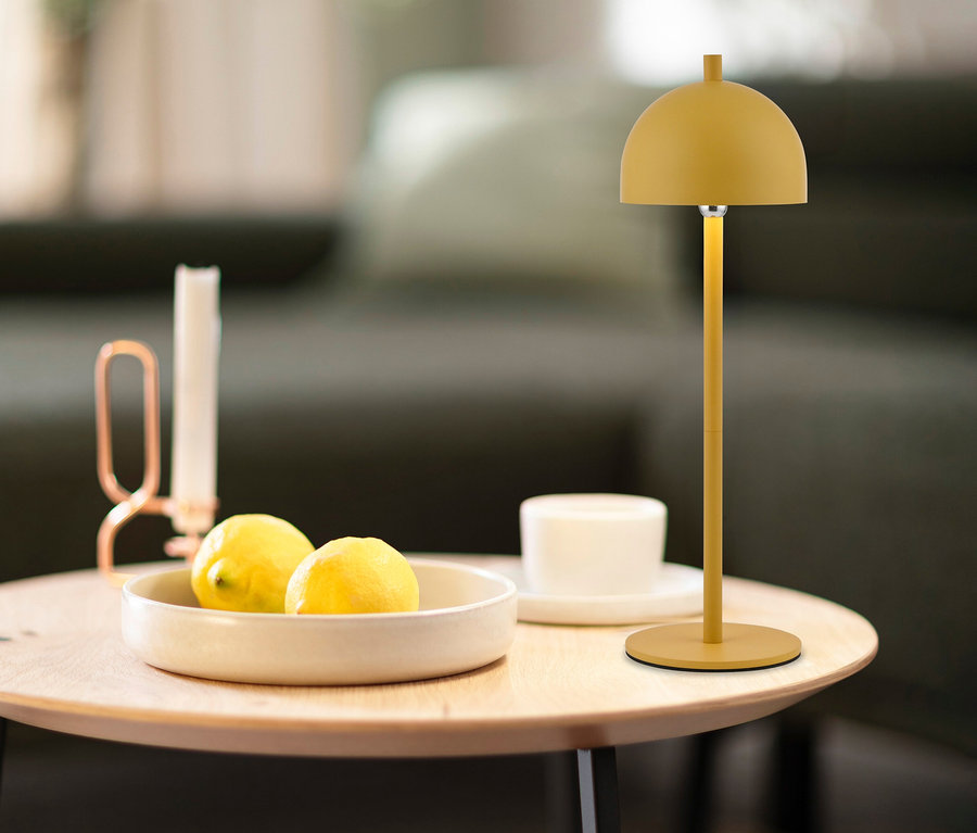 Gelbe SCHÖNER WOHNEN - Kollektion Akku-Tischleuchte »Bellis« auf einem Holztisch, daneben eine Schale mit zwei Zitronen, eine Kerze und ein weißer Becher.