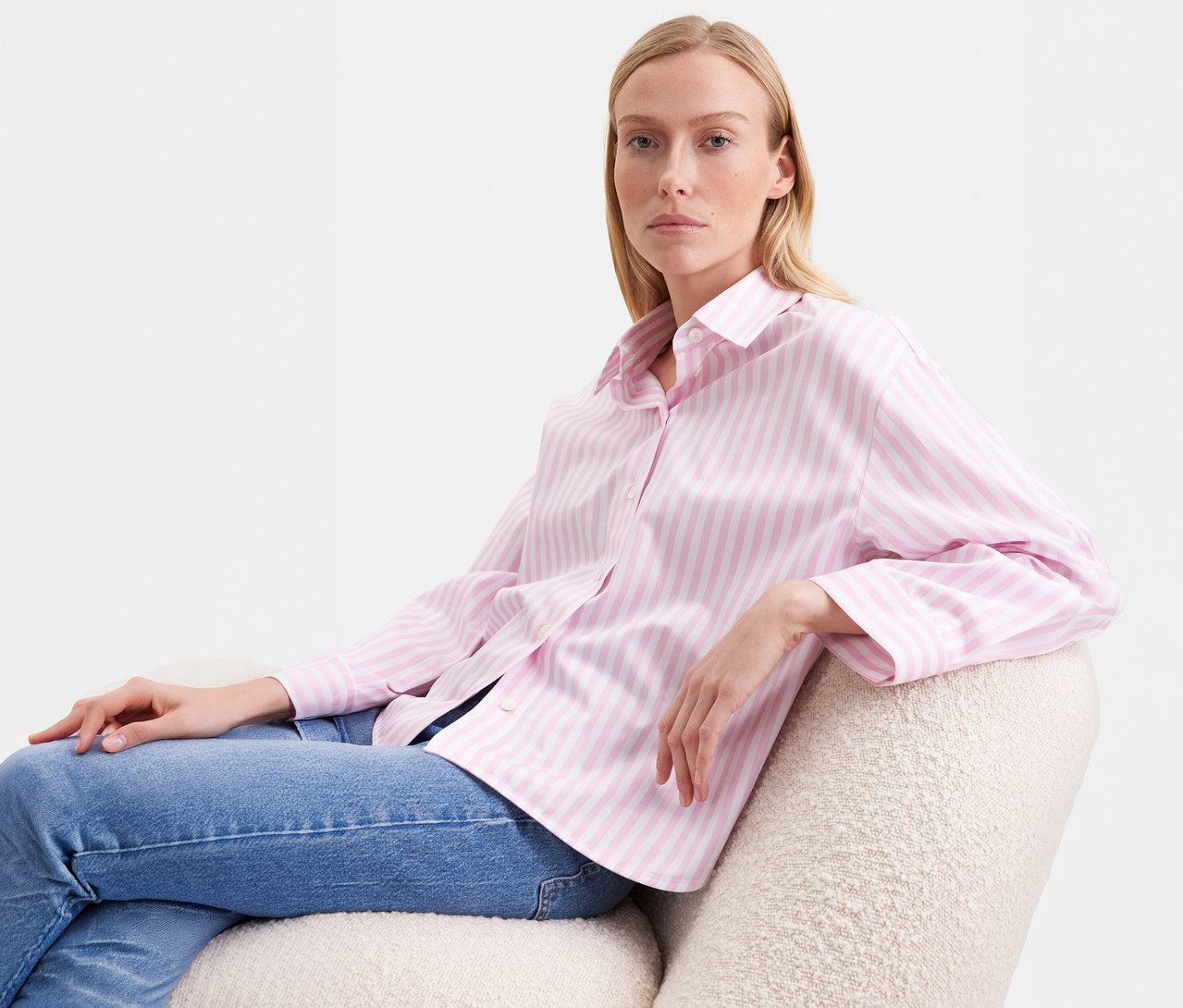 Blonde Frau sitzt in einem hellen Sessel und trägt eine roséfarbene Seidensticker Oversize Hemdbluse mit Streifen und blaue Jeans.