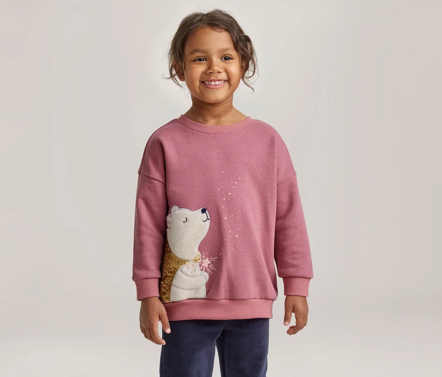 Ein lächelndes kleines Kind mit dunklen Locken trägt ein rosa Sweatshirt mit einem weißen Igel, dessen Körper goldfarben und strukturiert ist und der zu weißen Glitzerpartikeln aufblickt. Das Kind trägt auch dunkelblaue Hosen.