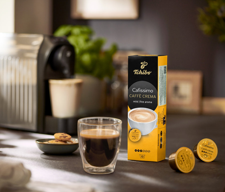 Auf einem Tisch stehen eine Kaffeemaschine, ein Glas Kaffee und eine Packung Cafissimo Caffè Crema mild - 120 Kapseln.