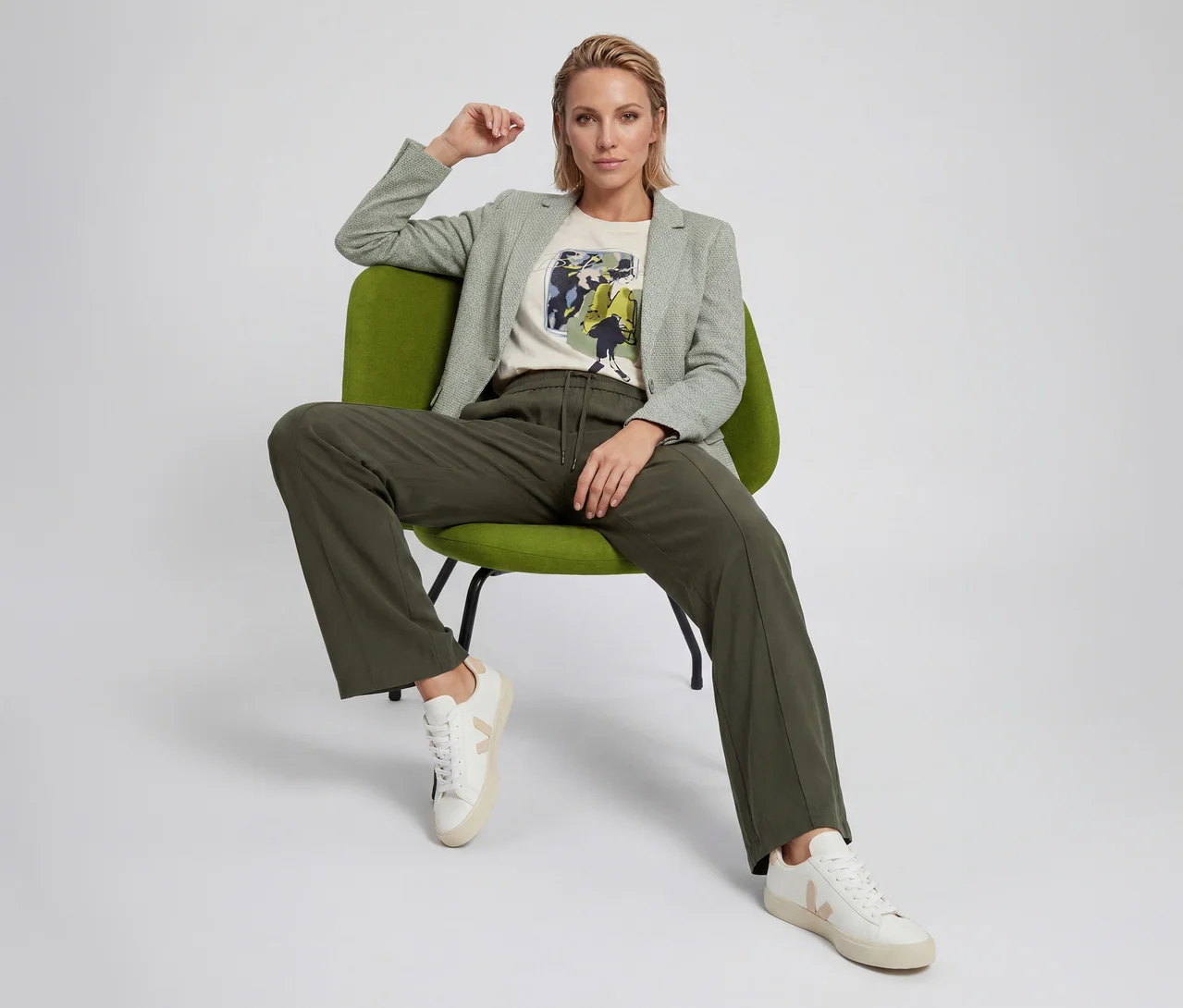 Frau sitzt auf einem grünen Stuhl, trägt einen hellgrünen gemusterten Blazer, ein cremefarbenes Grafik-T-Shirt, dunkelgrüne Hose mit Kordelzug und weiße Sneaker mit beigen V-Logo-Akzenten.
