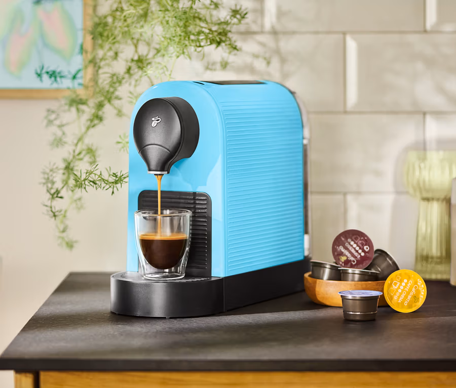 Eine türkisfarbene Kaffeemaschine füllt eine Tasse mit Kaffee. Kaffeekapseln liegen in einer Holzschale daneben.