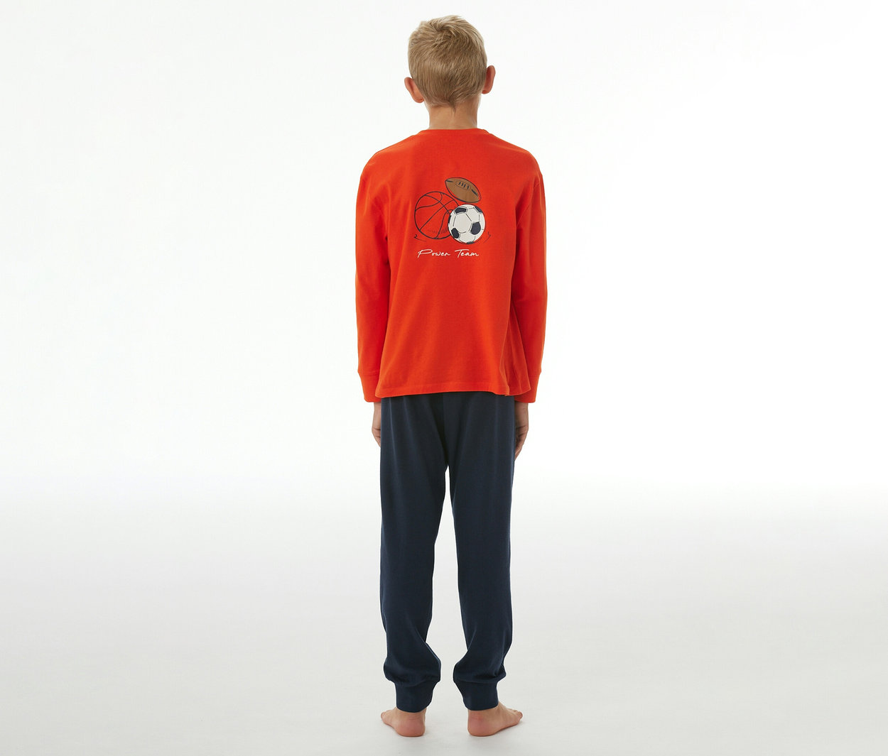 Ein Junge steht mit dem Rücken zur Kamera und trägt einen SCHIESSER Kinder-Pyjama mit Bündchen. Das orange Oberteil hat einen Aufdruck mit einem Basketball, Fußball und American Football sowie dem Schriftzug „Power Team“. Die Hose ist dunkelblau.