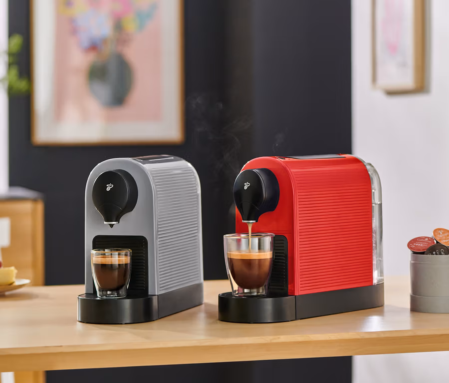 Zwei Kaffeemaschinen, eine graue und eine rote, stehen auf einem Holztisch. Die rote Maschine brüht Kaffee in eine Glastasse, während die graue Maschine eine volle Tasse hat. Ein Behälter mit Kaffeekapseln und ein Stück Himbeerkuchen sind ebenfalls auf dem Tisch.