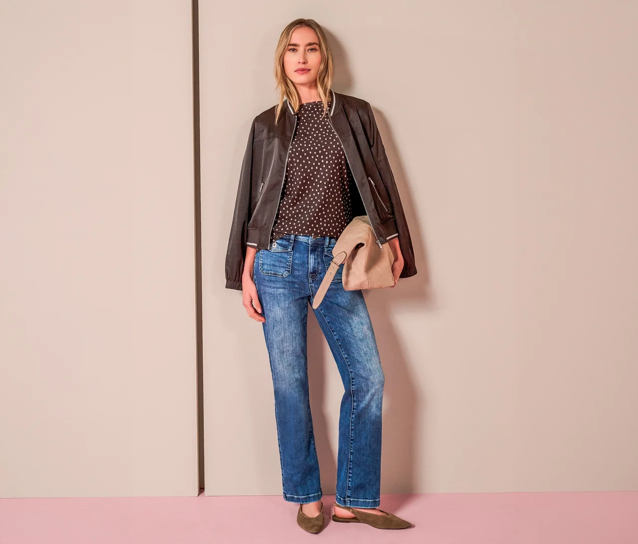 Eine Frau trägt ein gepunktetes Street One Blusenshirt, eine braune Bomberjacke, blaue Bootcut-Jeans und hält eine beigefarbene Tasche.