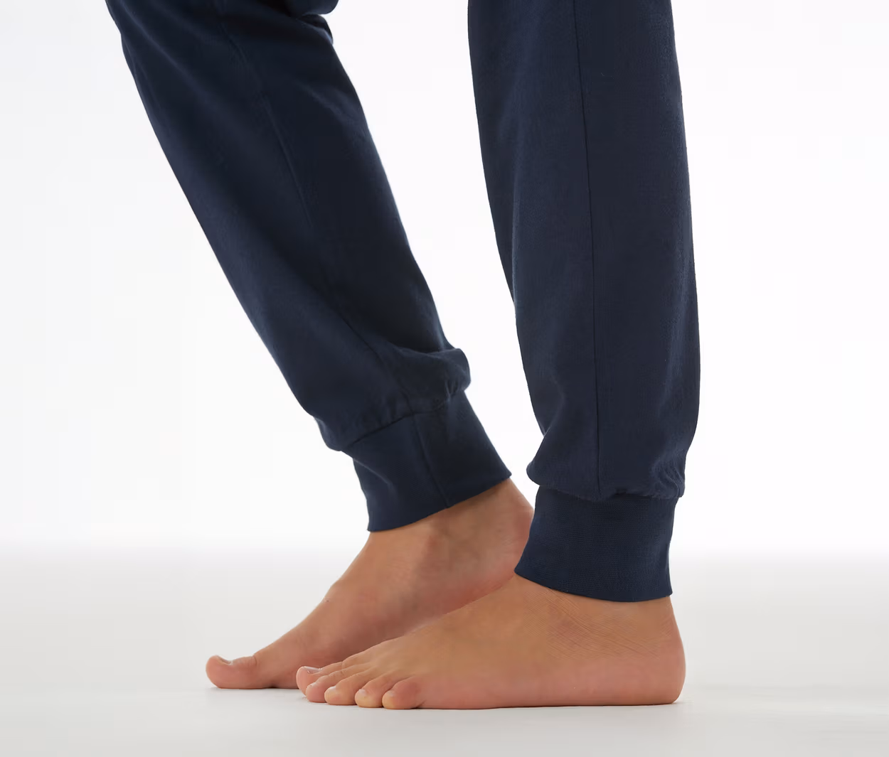 Unterschenkel und nackte Füße einer Person, die eine dunkelblaue SCHIESSER Kinder-Pyjamahose mit Bündchen trägt und auf einem weißen Untergrund steht.